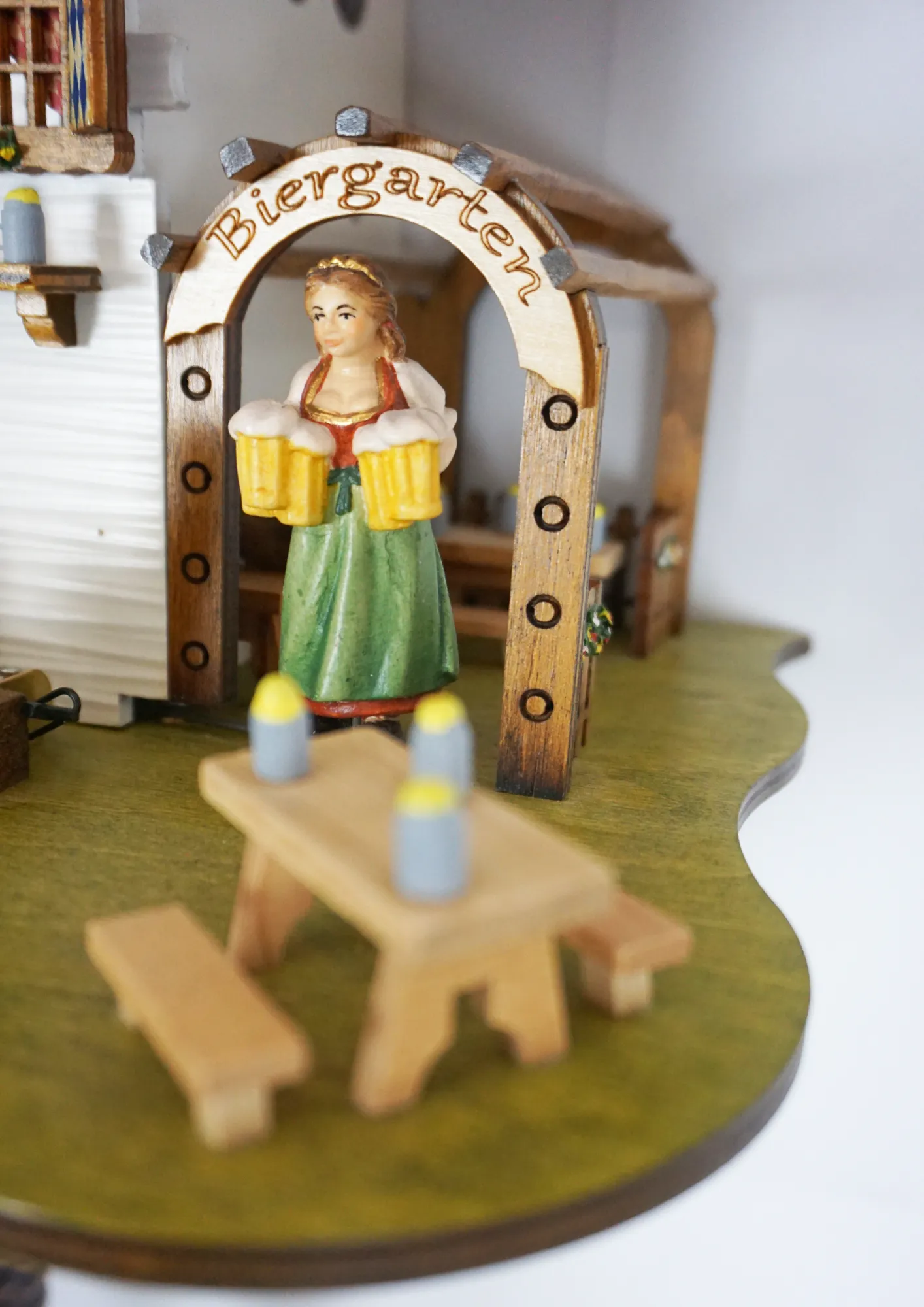 Cuckoo Clock  The Beer Garden  8 Day Movement with Music (4) Bavarian-style house to celebrate the infamous Oktoberfest. Blue and white shutters on the windows represent Bavaria. Around the clock, there are two men drinking beer and a lady wearing a dirndl dress, serving. The cuckoo calls every half hour and plays